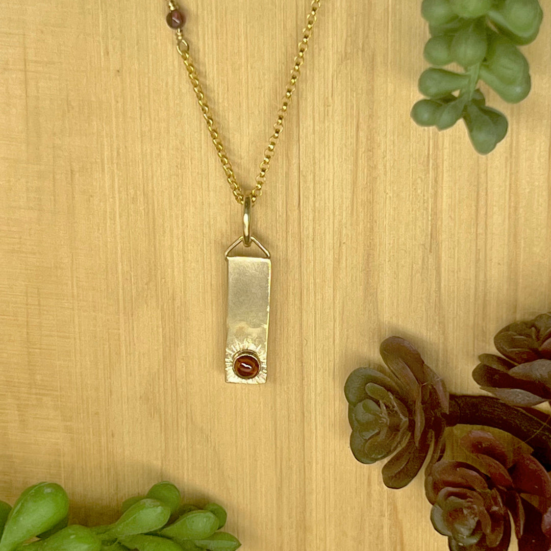 Gold bar necklace with garnet on wood background with foliage