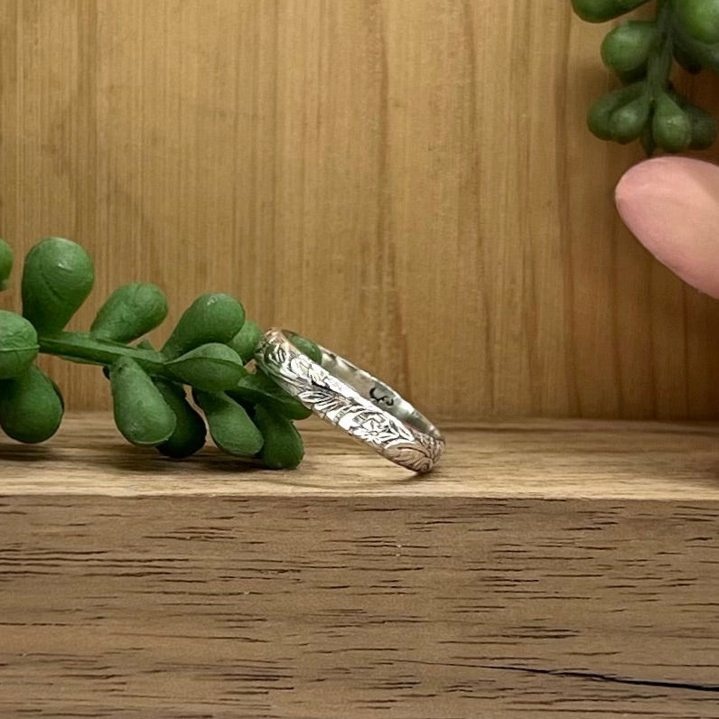 Silver ring with engraved design on a wooden surface with green leaves